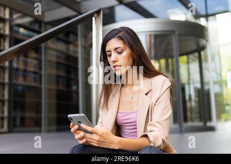 Focused young Middle Eastern female manager in blazer sitting near entrance of modern building and texting message on cellphone Stock Photo