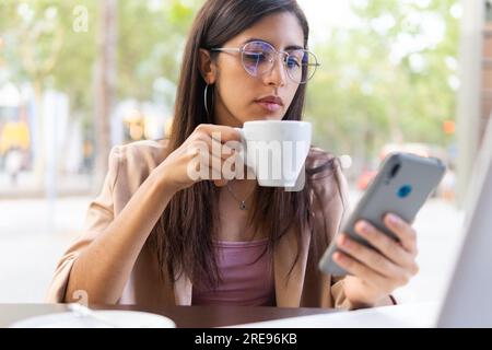 Pensive young Hispanic businesswoman in eyeglasses drinking coffee and browsing on smartphone while looking at screen Stock Photo