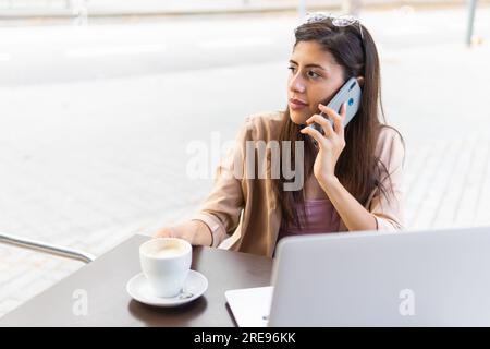 From above of young Hispanic female entrepreneur sitting at table with coffee near laptop and talking on smartphone while looking away Stock Photo