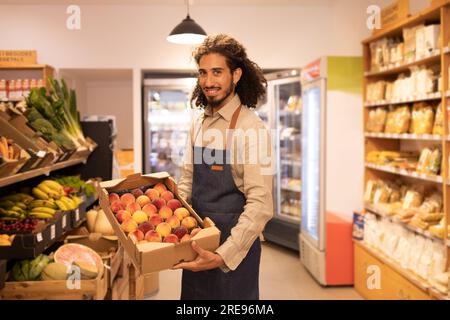 Side view of positive young bearded ethnic male seller with long curly hair in apron smiling and looking at camera while demonstrating box with fresh Stock Photo