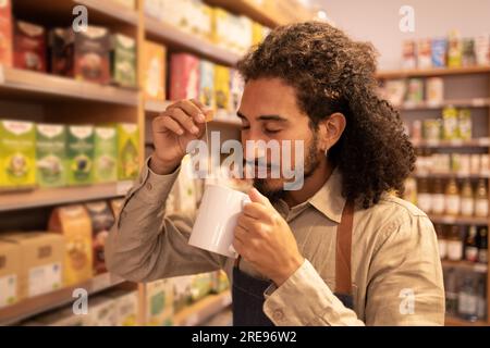 Portrait of serious curly hair male worker drinking hot tea from white mug while standing near shelves with tea box good in modern grocery shop Stock Photo