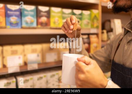 Cropped unrecognizable male worker drinking hot tea from white mug while standing near shelves with box of tea good in modern grocery shop Stock Photo