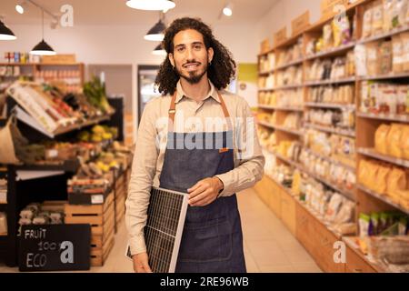 Positive seller male in casual clothes and apron smiling and looking at camera while standing near shelves holding portable solar panel in modern supe Stock Photo