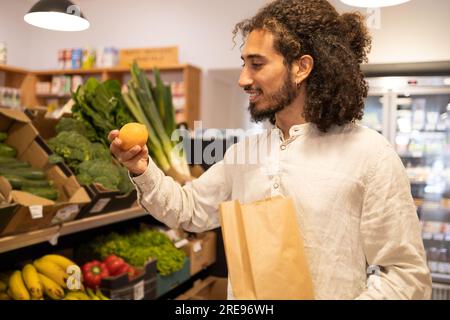 Portrait of young ethnic man with curly hair in casual clothes standing near shelves with assorted fresh vegetables and fruits while shopping indoors Stock Photo
