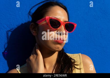 Portrait of serious young Indian female holding sunglasses and looking at camera while standing in light with shadow in blue background wall Stock Photo