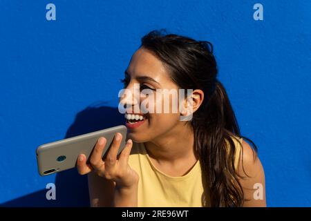 Portrait of smiling young Indian female with long dark hair looking away while recording voice message against blue wall in bright sunlight Stock Photo