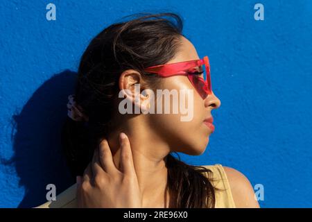 Portrait of young Indian female holding sunglasses and looking away while standing in light with shadow in blue background wall Stock Photo
