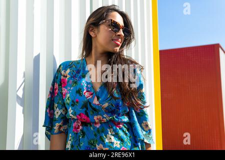 From below confident young Indian female in sunglasses looking away with long dark hair and standing in daylight against gray striped wall in daylight Stock Photo