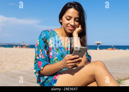 Portrait of smiling young Indian female with long dark hair looking at screen of mobile phone while sitting on beach with hand near face in daylight Stock Photo