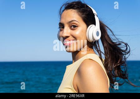 Side view of happy young Indian female listening to music in wireless headphones while resting on rocky coast near waving sea Stock Photo