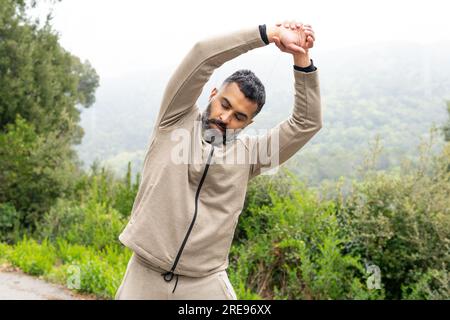 Young bearded Indian male in sportswear standing on road with closed eyes tilted head holding hands above head while doing stretching exercise in day Stock Photo