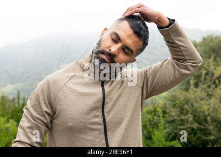 Young bearded Indian male in sportwear with closed eyes touching tilted head while standing and exercising in green meadow on sunny day in countryside Stock Photo