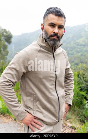 Serious bearded Indian male athlete in sportswear TWS earbuds looking at camera while standing with hands on waist against blurred green trees Stock Photo
