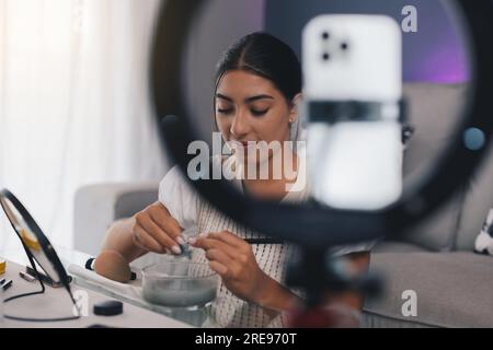 Young positive female blogger adding clay powder into water making skincare product while shooting video on cellphone at home Stock Photo