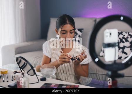 Young Hispanic female influencer applying bronzer with brush while recording makeup tutorial on smartphone in modern room Stock Photo