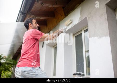 Low angle side view of bearded adult male painter in casual clothes looking away while standing near window and painting cement wall under roof with r Stock Photo