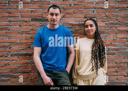 Smiling man in blue t shirt and Hispanic woman with braided hair in beige clothes standing near brick wall and looking at camera Stock Photo