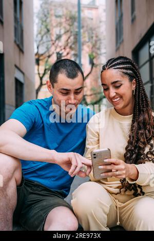 Smiling man in blue t shirt and Hispanic woman in beige outfit sitting on street and using cellphone together Stock Photo