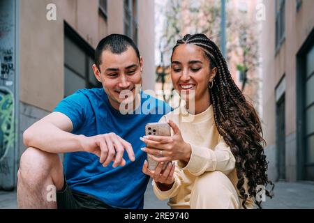Smiling man in blue t shirt and Hispanic woman in beige outfit sitting on street and using cellphone together Stock Photo