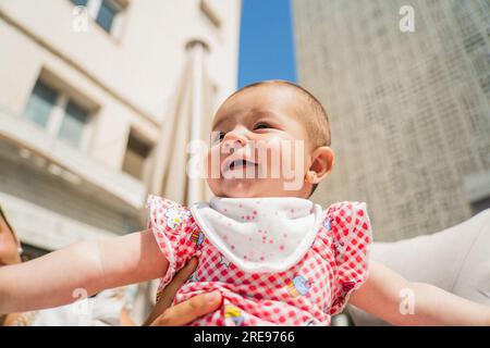 From below of cute little baby in casual clothes smiling and looking away while carried by crop anonymous parent against modern building background on Stock Photo