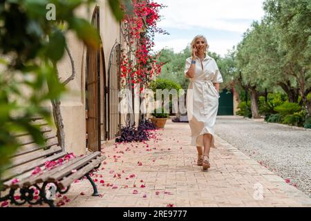 Smiling elderly female with gray hair and with hand in pocket looking at camera while walking on sidewalk with plants bench building with fence doors Stock Photo