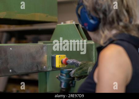 Unrecognizable female in earmuffs and gloves looking down while standing near heavy machine with yellow box and pressing red stop button in daylight d Stock Photo