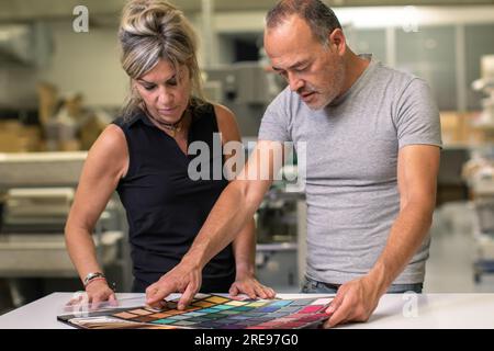 Serious mature male and adult female in casual clothes standing near table while examining various colorful fabric samples for quality and textures in Stock Photo