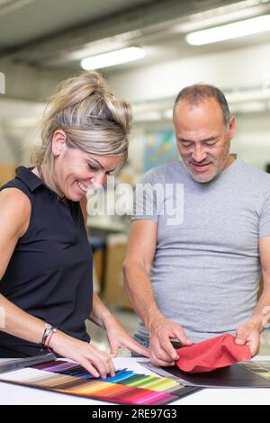 Smiling mature male and adult female in casual clothes looking down while standing near table and examining various colorful fabric samples for qualit Stock Photo