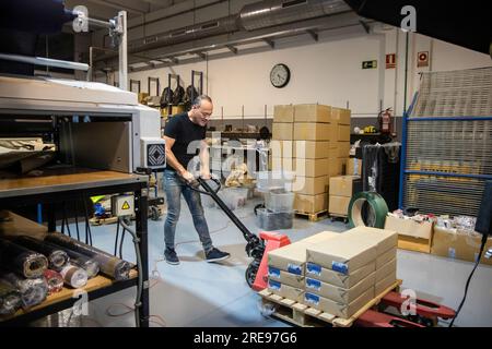 Side view of mature male worker looking down while standing and pulling packed material on pallet jack in production factory against carton boxes and Stock Photo