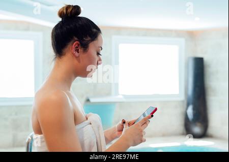 Side view portrait of focused young female in white towel standing and browsing mobile phone in bright indoor swimming pool at daylight Stock Photo