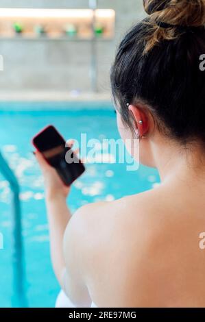 Back view of unrecognizable female in white towel using smartphone while sitting on swimming pool terrace during summer vacation Stock Photo