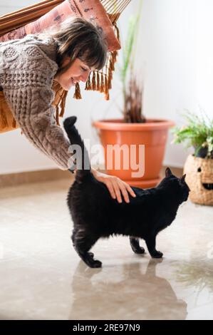 Side view of positive young female in casual sweater lying on hammock bed while stroking cute black Scottish fold cat over smooth room floor in modern Stock Photo