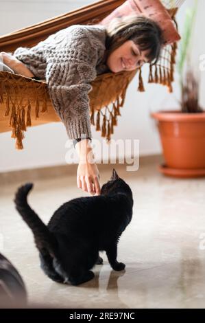Side view of positive young female in casual sweater lying on hammock bed while stroking cute black Scottish fold cat over smooth room floor in modern Stock Photo
