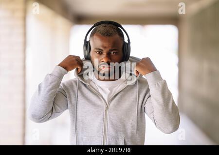 Portrait of young African American bearded male in sportswear looking at camera while standing with the headphones in your ear and touching hood of ja Stock Photo