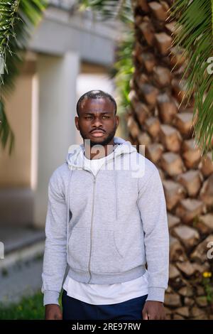 Thoughtful young African American male athlete in sportswear looking away while standing under green leaves palm tree against blurred building with pi Stock Photo