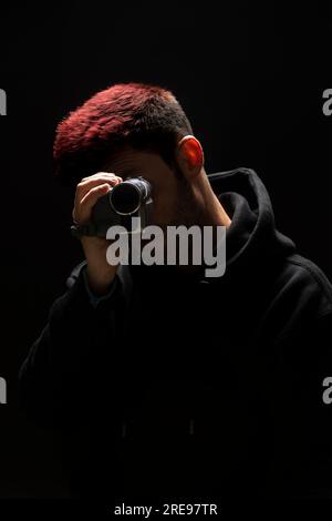 Portrait of young unrecognizable male in cozy clothes with red dyed hair recording video on portable camera while standing against black background Stock Photo