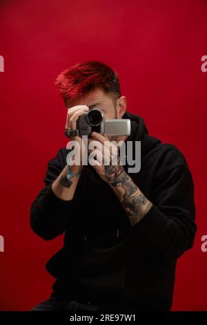 Portrait of anonymous young male in cozy clothes with red dyed hair recording video on portable camera while standing against red background Stock Photo