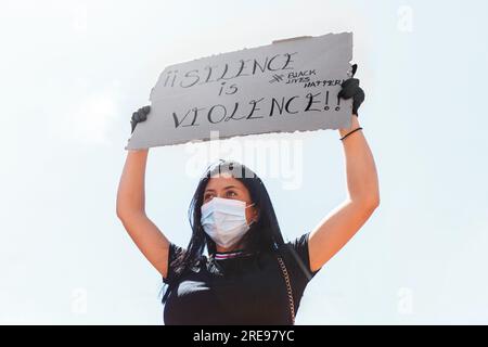 Low angle of female wearing medical mask and gloves standing with raised arms and carton placard with silence is violence and back lives matter inscri Stock Photo