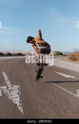 Full body young bearded skater in casual outfit jumping while performing kickflip on skateboard on asphalt road Stock Photo