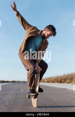 Full body young bearded male skater standing on edge of skateboard keeping balance while performing trick on asphalt road with hand raised and looking Stock Photo