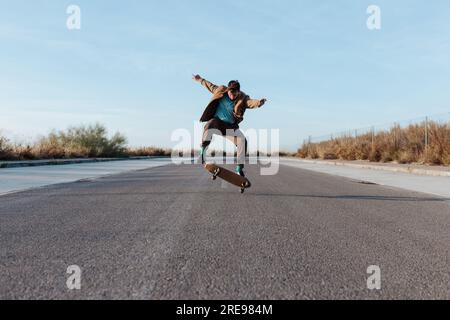 Full body young bearded skater in casual outfit jumping while performing kickflip on skateboard on asphalt road Stock Photo