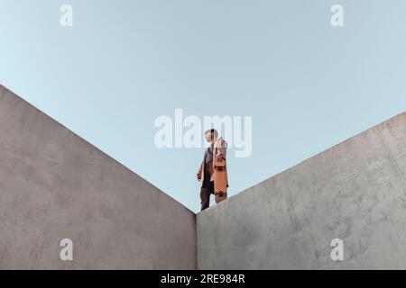 From below of young bearded male skater standing on high concrete ramp in skatepark with skateboard looking away Stock Photo