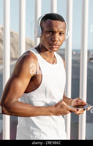 Close up of African American runner browsing mobile phone and listening to music in headphones while looking at camera training on street Stock Photo