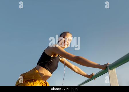 Young cheerful athletic caucasian woman leaning on banister outdoors and looking at camera after working out Stock Photo