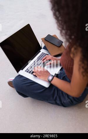 African American self employed woman sitting on street with laptop and browsing smartphone Stock Photo