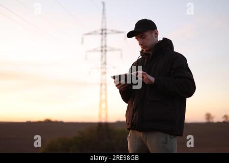 Young power engineer holds digital tablet against high voltage tower at field. Electrical engineer checking energy efficiency electrical towers. World Stock Photo
