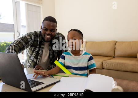 Happy african american father and son sitting table using laptop and doing schoolwork together Stock Photo