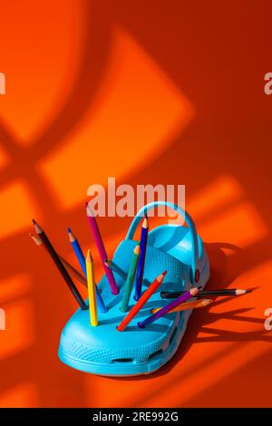 High angle of colorful pencils inserted into the holes of bright blue plastic clogs placed on orange background during daytime Stock Photo