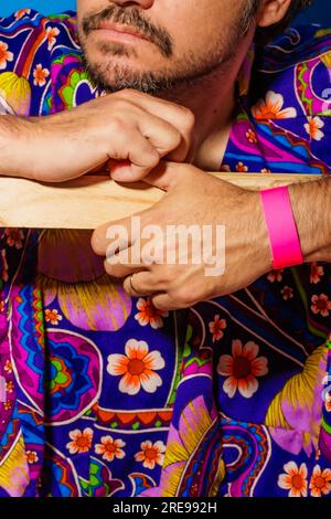 Crop bearded male in multicolored vibrant shirt with ornamental floral pattern and pink resort wristband leaning on wooden plank Stock Photo