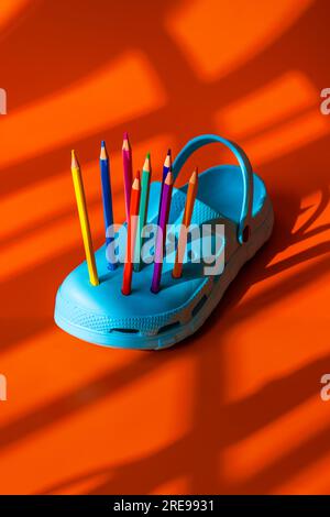 Colorful pencils inserted into the holes of bright blue plastic clogs placed on orange background during daytime Stock Photo